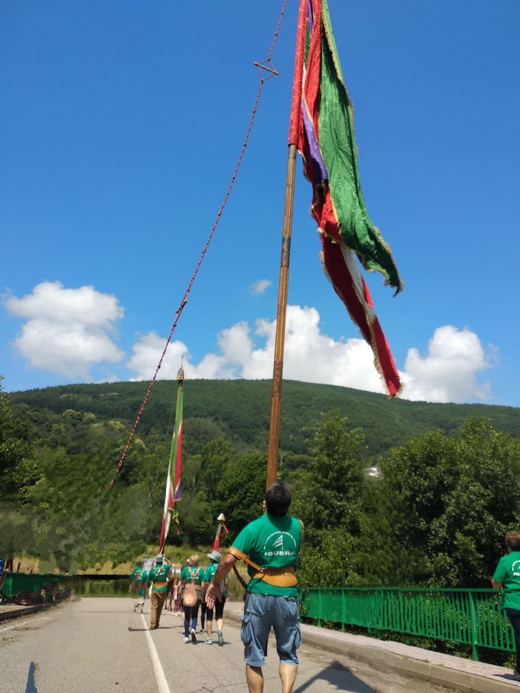 Un pendonero con camiseta verde de Igüeña lleva el pendón a cuestas por una carretera, seguido de otros pendones y de gente andando, con el monte al fondo.