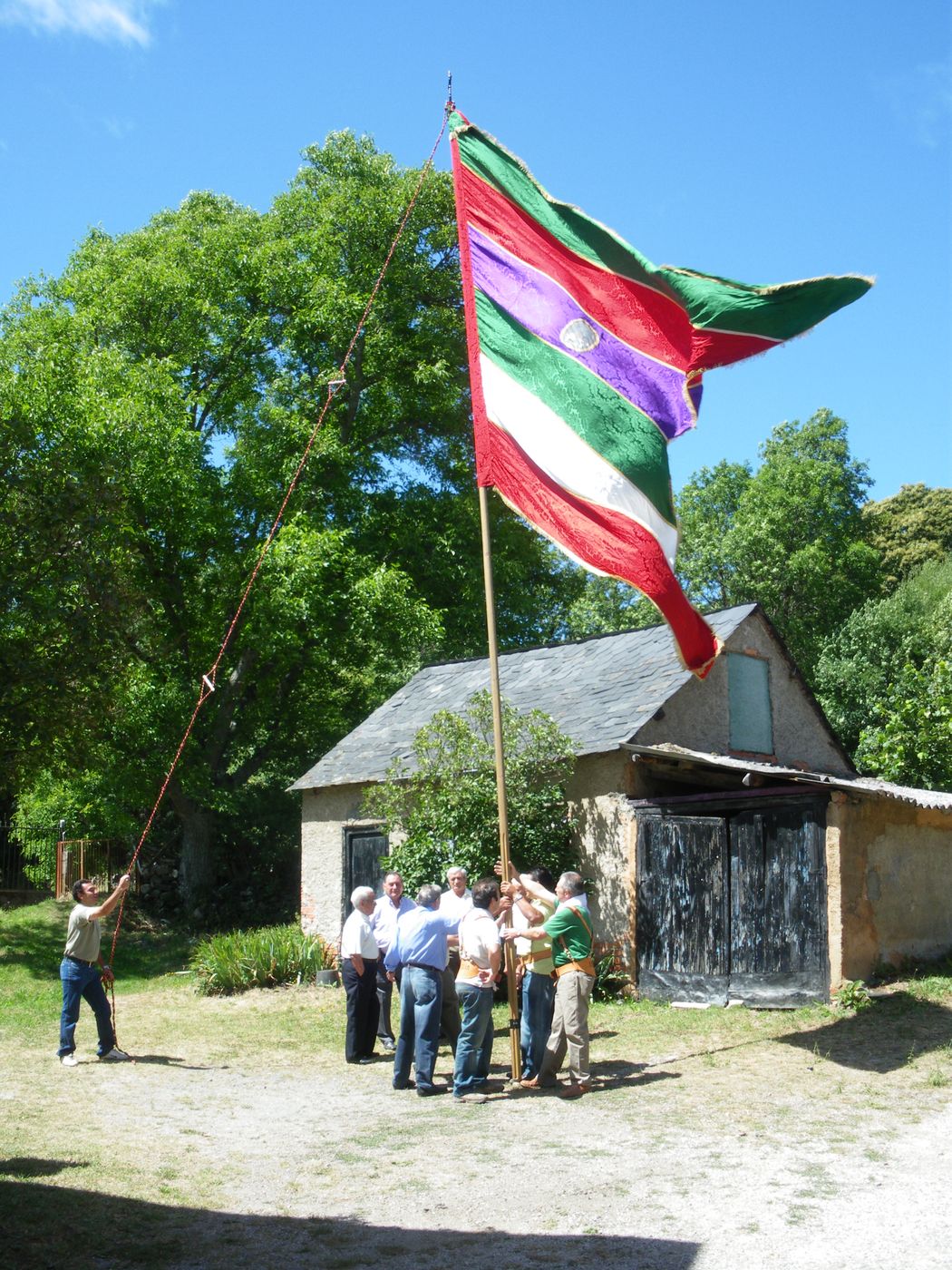 Varios hombres levantan el pendón de Igüeña junto a una casa de piedra con tejado de pizarra; el paño de franjas verdes, rojas, blancas y moradas ondea contra el cielo azul.